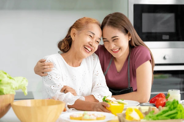 An adult daughter in a maroon top and apron laughing with her mother in a white top while cooking together in a bright kitchen