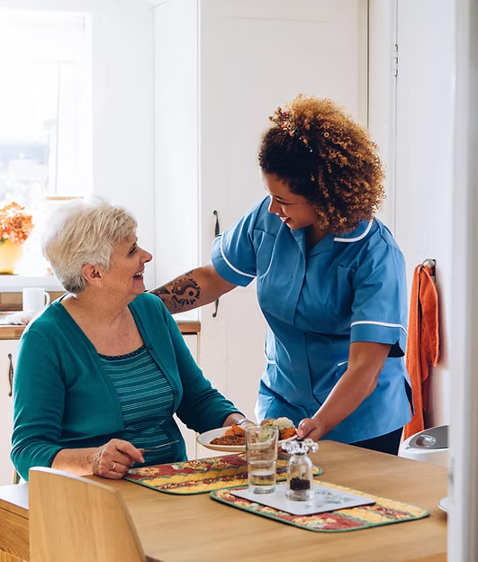 A caregiver in blue scrubs with curly hair smiling while serving a plated meal to a seated older woman in a teal cardigan