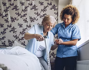 A caregiver in blue scrubs helping an older woman in gray stand up from a bed using a cane